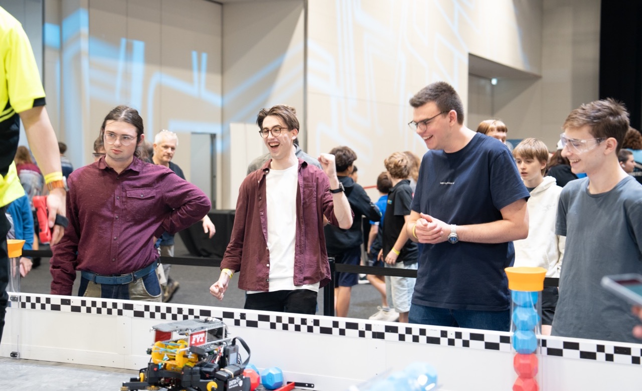 Four club members cheering next to and looking at a VEX Robotics field.